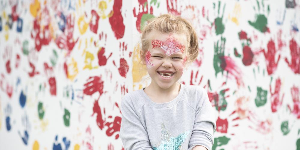 A small blonde girl with face paints smiles. The background is a white wall with painted hand prints on.